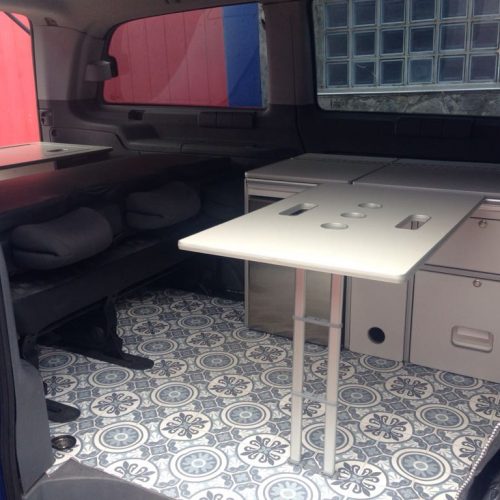 Interior de una furgoneta camper con suelo de baldosa decorativa, mesa plegable y área de asientos.