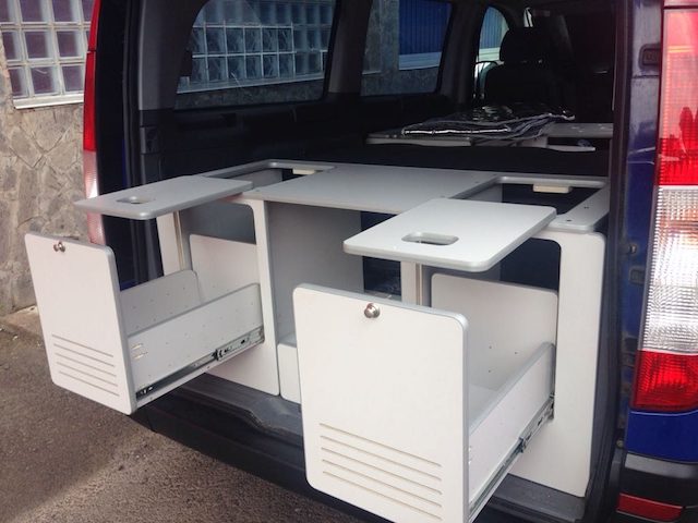 Mueble de almacenamiento multifuncional con mesas abatibles en la parte trasera de una furgoneta camper.