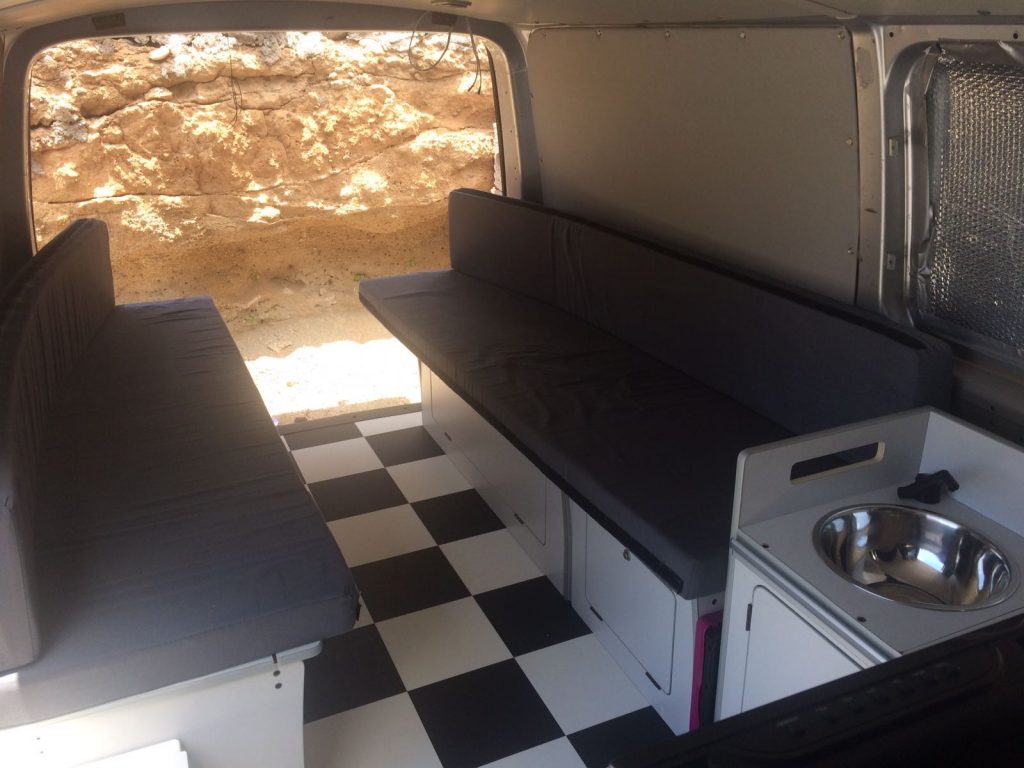 Interior de una furgoneta camper con área de asientos, suelo de tablero de ajedrez y cocina incorporada.