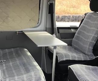 Interior de una furgoneta camper con asientos de tela a rayas y una mesa plegable gris.