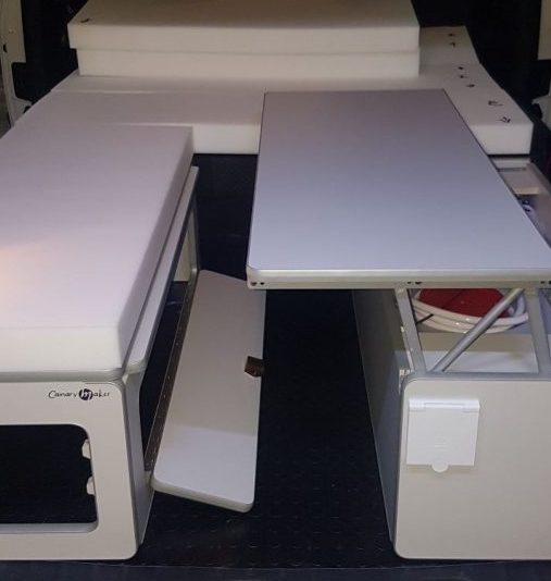Mueble camper multifuncional con secciones desplegadas en el interior de una furgoneta, incluyendo una mesa y espacios de almacenamiento.