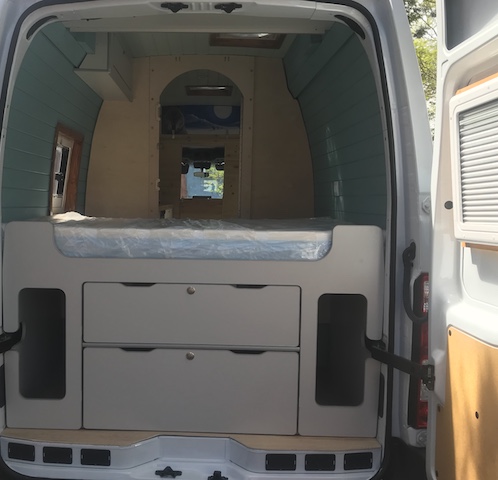 Vista trasera de una furgoneta camper durante el proceso de camperización, mostrando una cama con colchón protegido por plástico y muebles de almacenamiento.