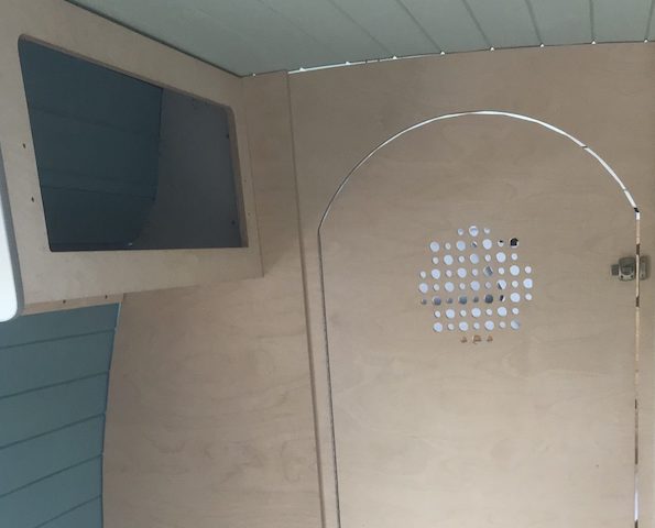 Interior de una furgoneta camper con paneles de madera y una puerta decorativa con patrón circular de agujeros.