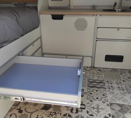 Cajón de almacenamiento abierto en una furgoneta camper con interior de cocina y suelo estampado.