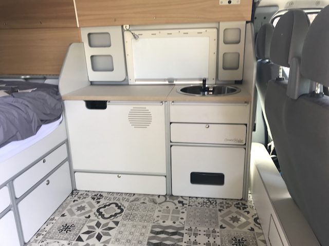Interior de furgoneta camper con zona de cocina que incluye fregadero y armarios, sobre suelo de baldosas de patrón.