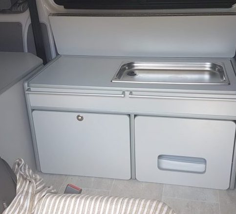 Cocina de furgoneta camper equipada con fregadero y almacenamiento debajo, junto a un asiento con cojín a rayas.