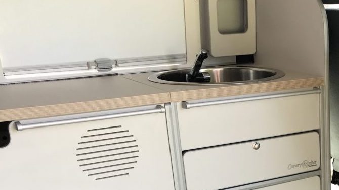 Área de cocina en una furgoneta camper con un fregadero de acero inoxidable y encimera blanca.