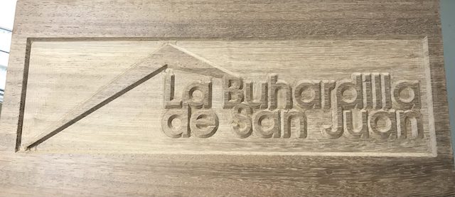 Talla en madera con el nombre 'La Buhardilla de San Juan' dentro de un contorno de casa estilizada.
