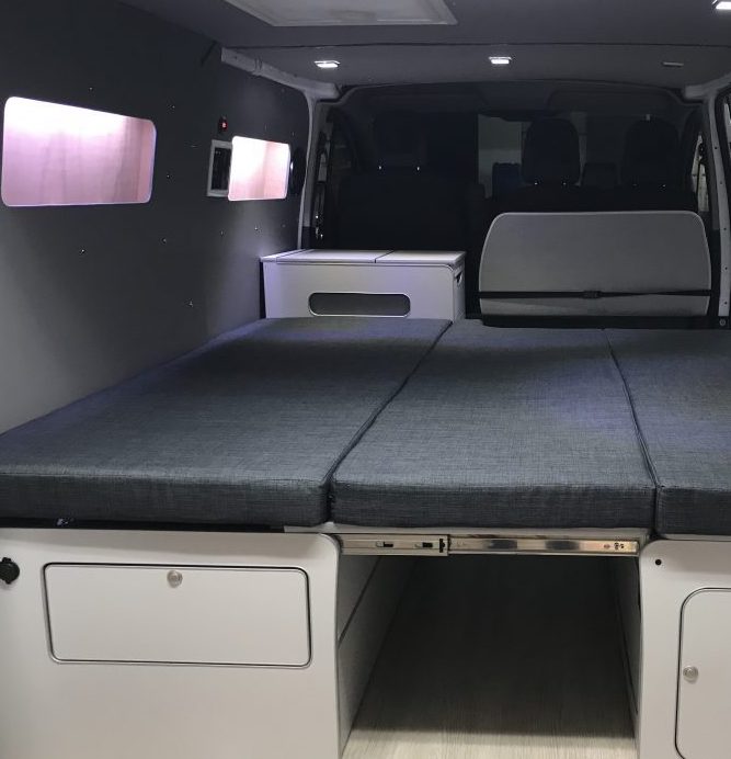 Interior de una furgoneta camper Ford Transit Custom con cama desplegada y almacenamiento debajo.