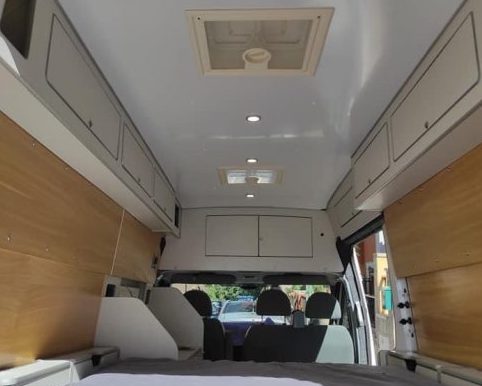 Vista interior de una furgoneta camperizada con una cama grande, armarios de almacenamiento y claraboya.